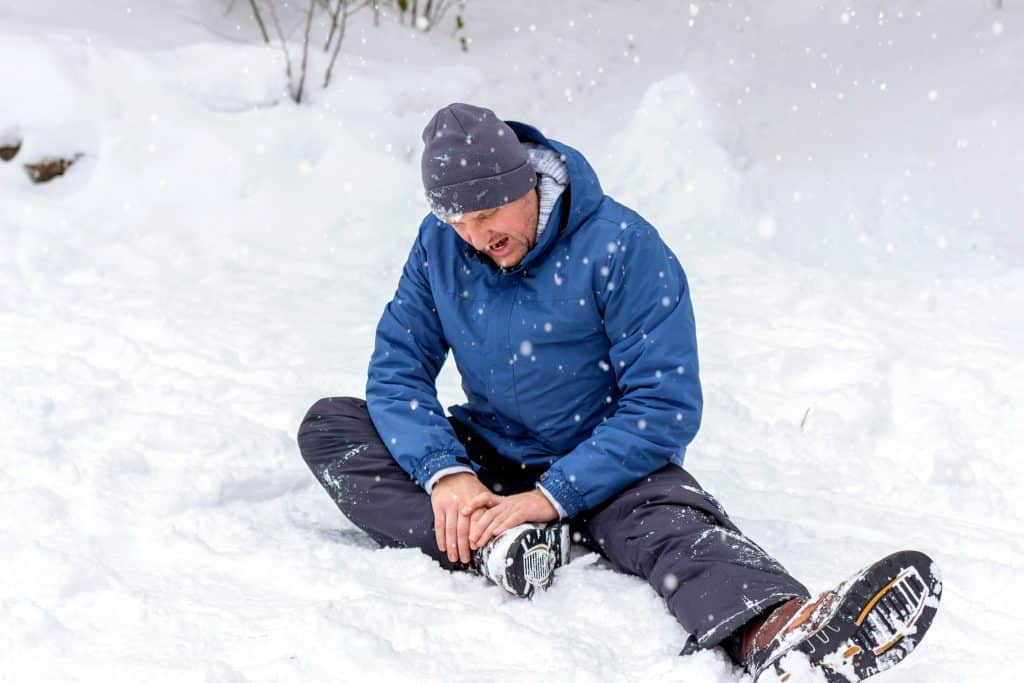 Unhappy young man with injured painful leg sitting on snowy road, outdoors. Portrait of Caucasian young male feeling pain in ankle after walking outside in snowy winter park. 